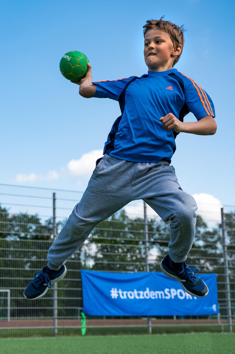 Junge springt auf einem Sportplatz und wirft einen grünen Handball, umgeben von einem Zaun und Bäumen im Hintergrund.