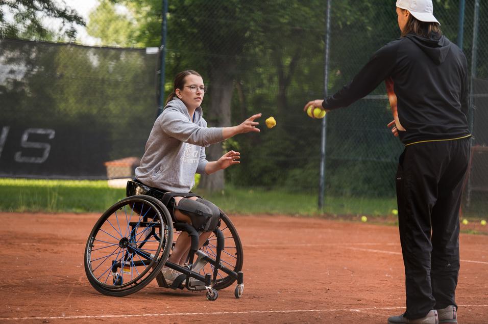 Eine Rollstuhlfahrerin fängt einen Tennisball von einem Trainer auf einem roten Tennisplatz.